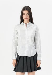 Striped button-up shirt in white with dark vertical lines, featuring a collar and long sleeves. Paired with a pleated dark skirt.