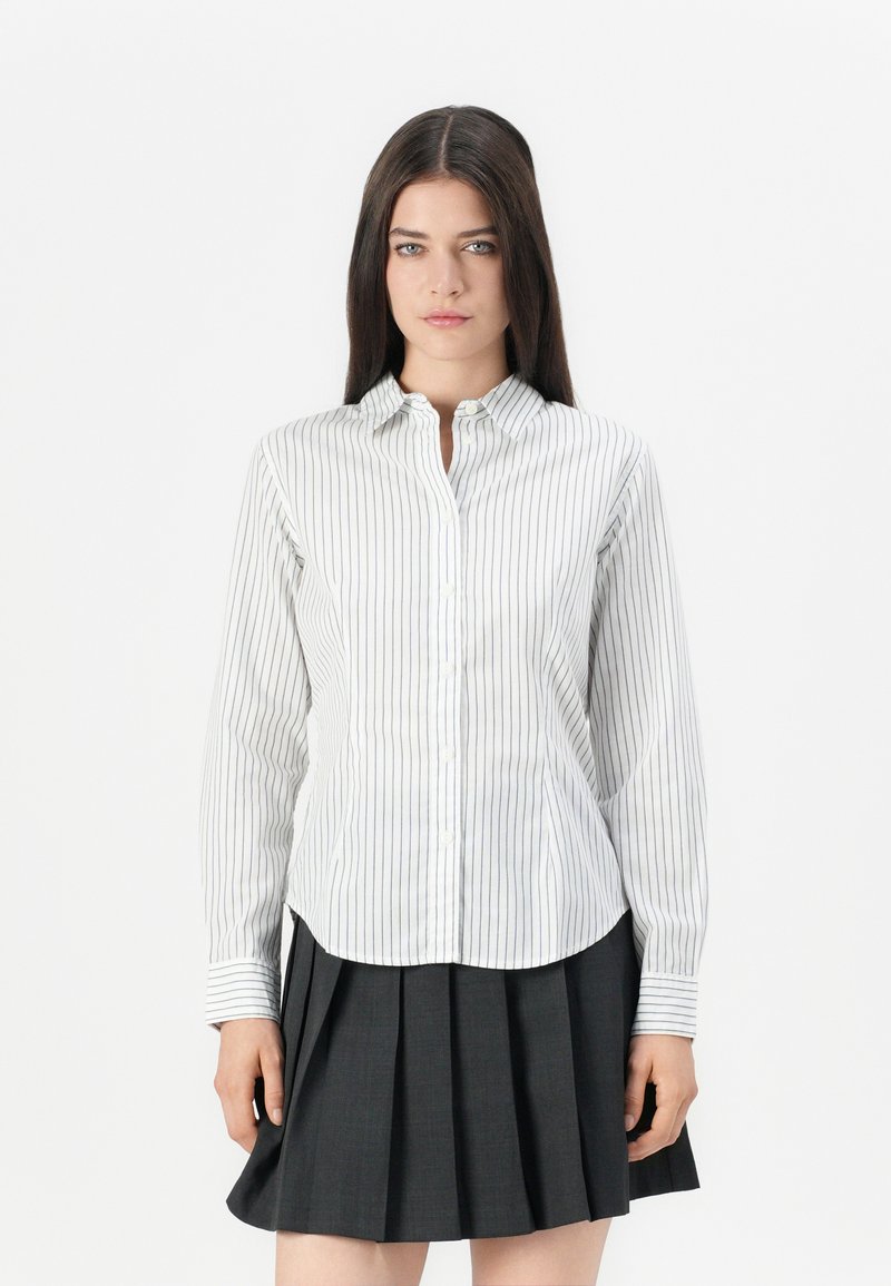 Striped button-up shirt in white with dark vertical lines, featuring a collar and long sleeves. Paired with a pleated dark skirt.
