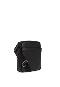 Borsa a tracolla in pelle nera con una tasca con zip frontale, cuciture rosse, tracolla regolabile e un dettaglio del logo. Forma rettangolare compatta.