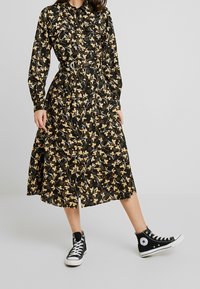 Nainen, joka on pukeutunut mustaan midi kukkamekkoon, jossa on pitkät hihat ja vyö, yhdistettynä mustiin korkeakorkoisiin Converse-tennarit.