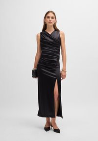 Black satin maxi dress with a draped design, asymmetrical neckline, side slit, and ruching details. Model holding a small black handbag.
