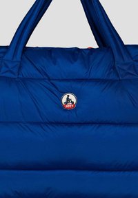 Borsa imbottita blu con doppie maniglie, caratterizzata da un logo Jott tondo. La texture è liscia con cuciture visibili e un motivo trapuntato.