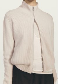 Cardigan zip-up rosa chiaro con texture a coste, collo alto e zip argentata. Indossato sopra un top neutro a strati, caratterizzato da un design semplice.