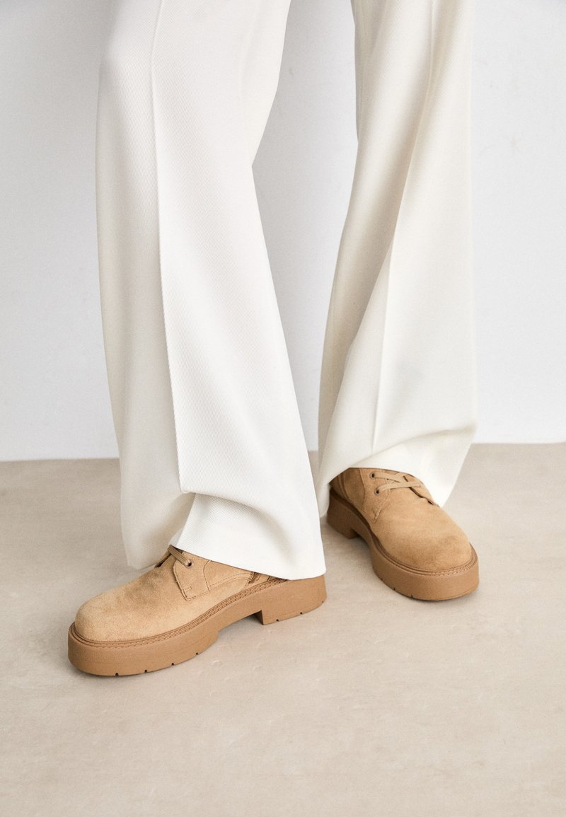 Kameljaskelised mokassiinid ümara toe, paeltega kinnituse ja paksu, tekstuuriga kummitald. Kombineeritud valgete, laiade jalgade pükstega.