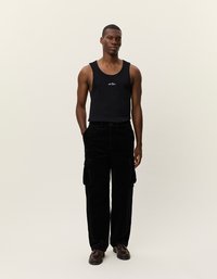 Schwarzes Tanktop mit weißem Logo, kombiniert mit schwarzen Cordhosen mit seitlichen Taschen. Dunkelbraune Schnürschuhe. Schlichter Hintergrund.