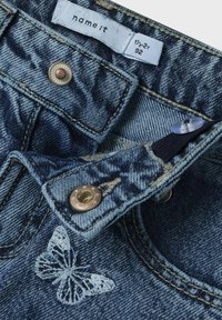 Jeans de mezclilla con un lavado azul, que presentan un bordado de mariposa, herrajes de botón de cobre y una etiqueta para la talla 1½-2 años.