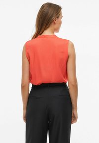 Blusa corallo senza maniche con una superficie liscia e scollatura tonda, abbinata a pantaloni neri a vita alta con tasche laterali.