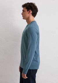 Niebieska koszula z długim rękawem, okrągłym dekoltem, miękką fakturą i subtelnym logo na piersi. Noszona z ciemnymi spodniami.