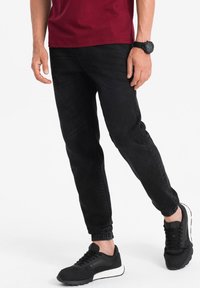 Svarta joggingsbyxor med elastiska muddar, tillverkade av mjukt tyg. I kombination med en vinröd t-shirt och svarta sneakers, vilket visar en avslappnad stil.