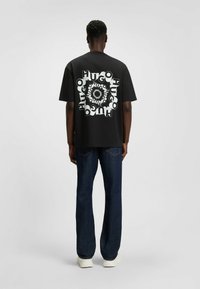 Sort kortærmet T-shirt med et gentagende hvidt "HUGO" tekstmønster på ryggen. Løs pasform, parret med mørkeblå jeans og hvide sneakers.
