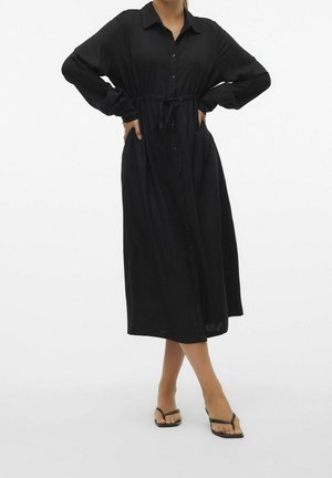 Femme portant une longue robe noire boutonnée avec une ceinture à la taille, debout les mains sur les hanches et portant des sandales noires ouvertes à l'avant.