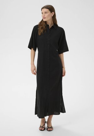 Frau trägt ein schwarzes Midi-Hemdkleid mit durchgehender Knopfleiste, kurzen Ärmeln und Seitenschlitzen, kombiniert mit schwarzen, Riemchen-Heeled-Sandalen.