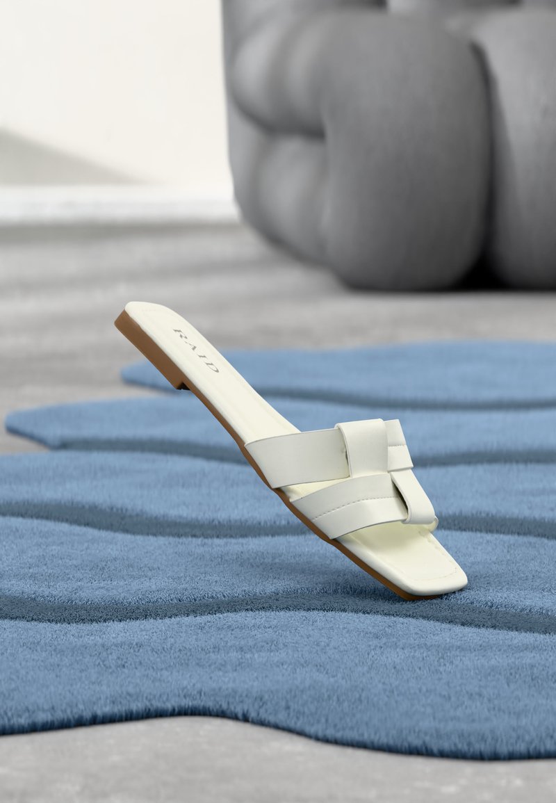 Sandale plateforme blanche à brides larges sur un tapis bleu texturé avec un oreiller gris surdimensionné flou en arrière-plan.