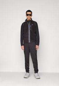 Man in een zwarte fleece vest over een langemouwen t-shirt, zwarte trainingsbroek, witte sneakers en zwarte wraparound zonnebril, staande tegen een neutrale achtergrond.