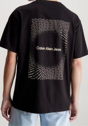 Personne portant un T-shirt noir avec le texte blanc « Calvin Klein Jeans » en motif circulaire dans le dos, associé à un jean bleu clair.
