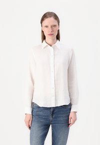 Camicia bianca in lino con colletto, maniche lunghe e orlo leggermente curvo. Abbinata a jeans in denim blu.