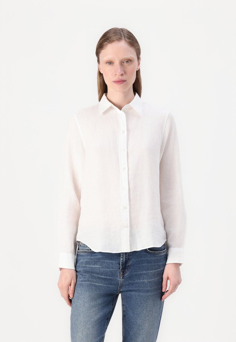 Camicia bianca in lino con colletto, maniche lunghe e orlo leggermente curvo. Abbinata a jeans in denim blu.