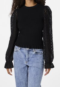 Mustat neulepaita, jossa on pitkät läpikuultavat pitsipuffihihat, joissa on leikattuja kuvioita ja röyhelöhelma, yhdistettynä vaaleansinisiin farkkuihin.