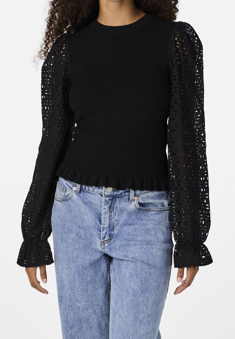 Mustat neulepaita, jossa on pitkät läpikuultavat pitsipuffihihat, joissa on leikattuja kuvioita ja röyhelöhelma, yhdistettynä vaaleansinisiin farkkuihin.