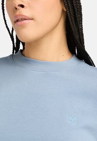 Felpa di colore azzurro chiaro con scollatura rotonda e colletto a costine, impreziosita da un piccolo logo bianco a forma di albero sul petto. Tessuto morbido e dal finitura strutturata.