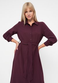 Langermet skjorte-kjole i dyp burgunder, med belte i livet og myk, strukturert stoff. Klassisk krage og oppbrettede ermer.
