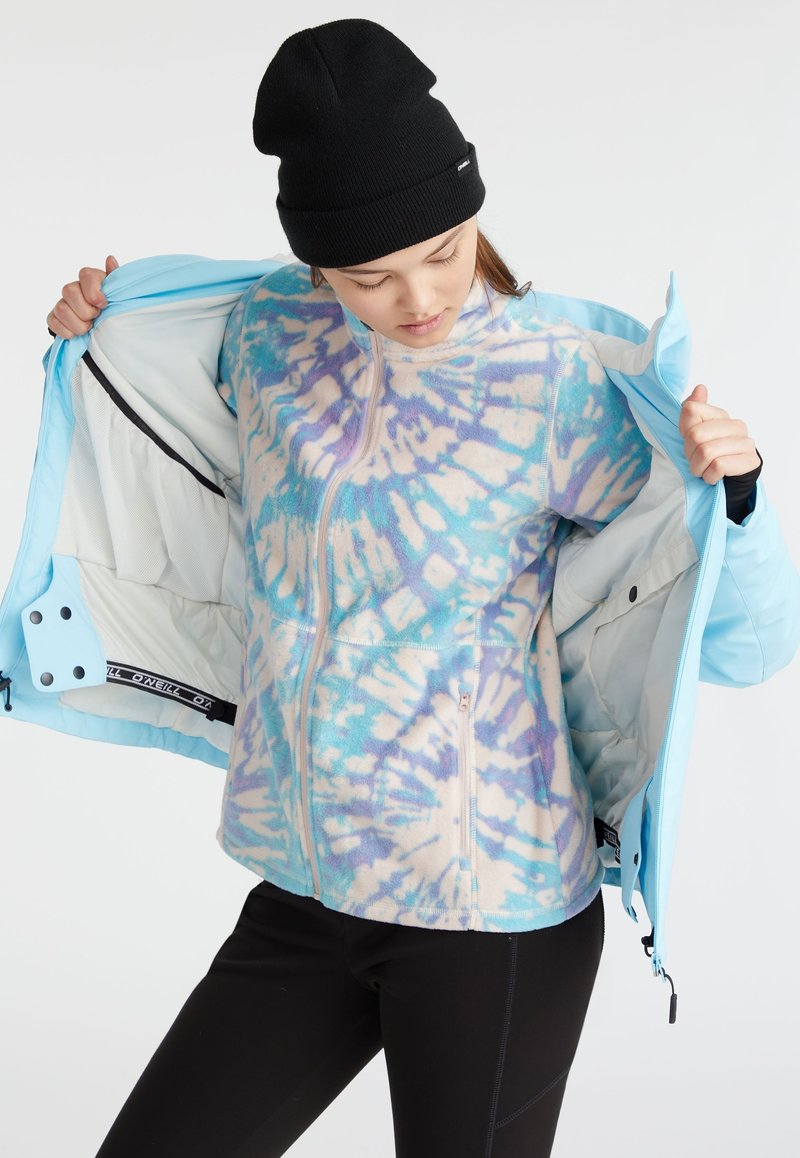 Giacca esterna azzurro chiaro con uno strato interno a motivo tie-dye nei colori pastello. Presenta una cerniera e una fodera interna in rete. Berretto nero indossato.
