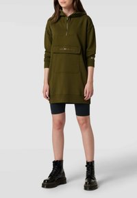 Oliwkowa sukienka-hoodie z zamkiem błyskawicznym z przodu, dużą kieszenią i logotypem; ma długie rękawy i luźny krój, zestawiona z czarnymi szortami rowerowymi.