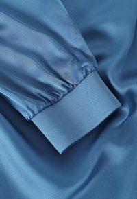 Tessuto di raso blu con una finitura lucida, caratterizzato da maniche arricciate e un polsino a costine a contrasto. Texture liscia e caduta morbida.