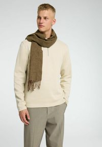 Top beige de manga larga con cuello de cremallera, combinado con una bufanda marrón a cuadros con flecos y pantalones de tela gris a medida.