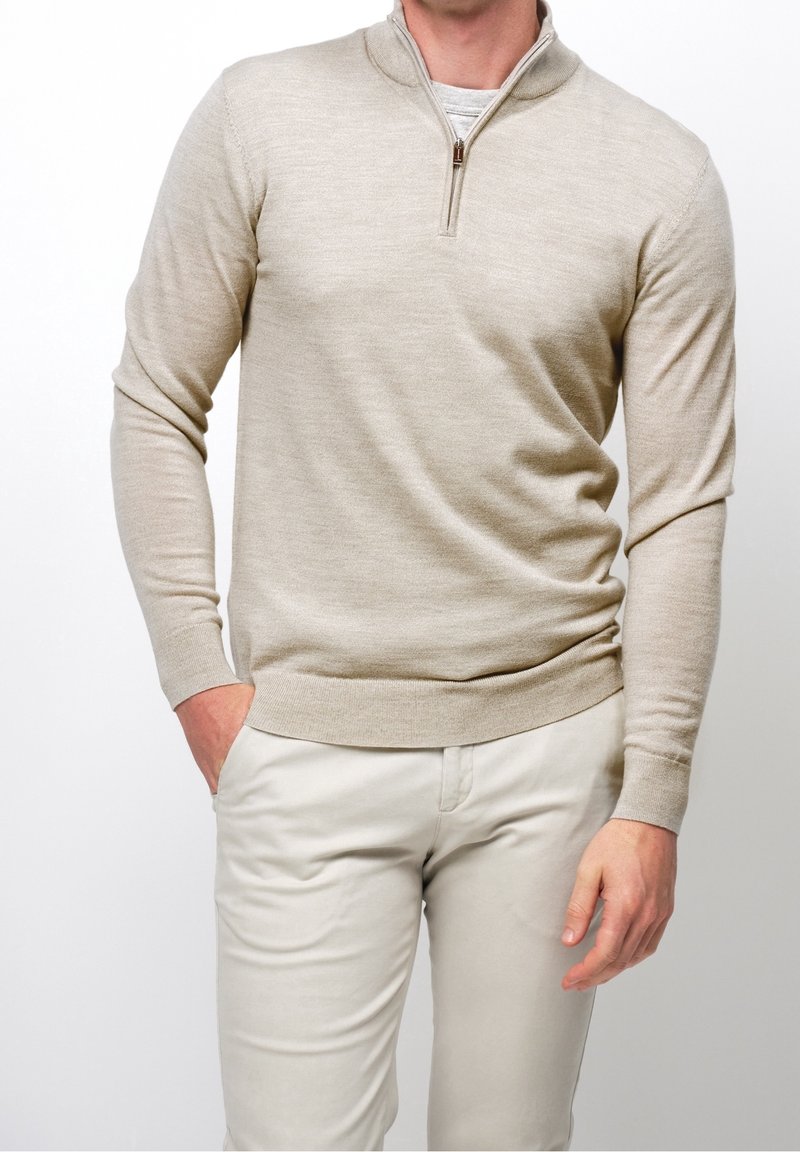 Homme portant un pull beige à demi-fermeture éclair et un pantalon beige clair, debout avec une main dans la poche, sur un fond blanc.