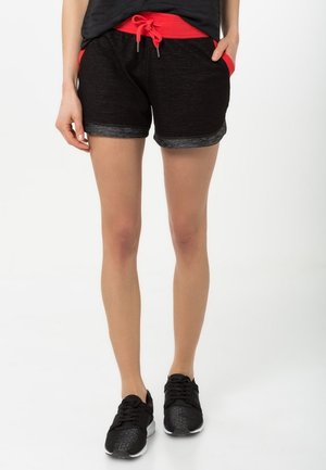 Frau trägt schwarze Sportshorts mit rotem Bund und Taschen, schwarze Sneakers, steht vor weißem Hintergrund.