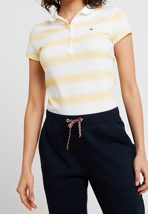 Vrouw draagt een geel-wit gestreept poloshirt met korte mouwen en zwarte broek met een rood-witte trekkoord.