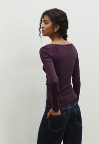 Top a maniche lunghe, aderente, di un profondo viola, realizzato in materiale elasticizzato con una scollatura ampia. Abbinato a jeans in denim blu scuro.