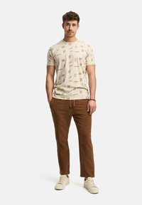 Beige T-Shirt mit kurzen Ärmeln und braunen geometrischen Mustern, kombiniert mit braunen strukturierten Hosen und hellen Sneakers. Einfach und lässig.