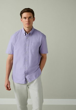 Mann steht vor einer schlichten Wand, trägt ein kurzärmliges lavendelfarbenes Hemd mit Knopfleiste und hellgraue Hose, schaut geradeaus mit neutralem Gesichtsausdruck.