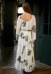 Langes, weißes Kleid mit schwarzen Blumenmustern, das lange Ärmel und einen runden Rückenausschnitt hat. Der Stoff wirkt leicht und fließend, in der Taille gerafft.