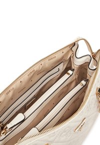 Valkoinen tikattu käsilaukku kultaoksilla. Sisäosassa on useita lokeroita ja beigellä vuorattu sisustus, jossa on toistuva "Guess" -kuvio.