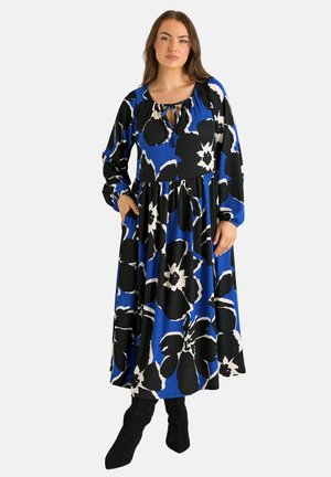 Femme portant une longue robe bleue à grand imprimé floral noir et blanc, manches longues, et bottes noires, debout avec une main dans la poche.