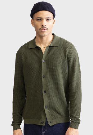 Olive green knit cardigan with a polo collar, button closure, textured pattern, and ribbed cuffs. Worn over a dark shirt and denim.