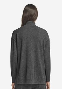 Person mit langen Haaren, die einen dunkelgrauen Rollkragenpullover und schwarze Jeans trägt, von hinten vor einem schlichten Hintergrund gezeigt.