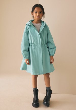 Ūdensizturīgs tirkīzzils mētelis ar kapuci, vidukļa auklu un elastīgām aprocēm. Valkāts pār pelēku topu, kombinēts ar melnām potīšu zābakiem un pelēkām zeķēm.