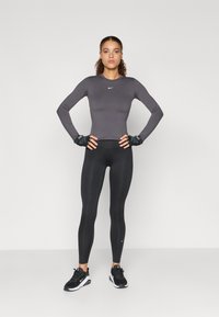 Siva dolgi športni top z belim logotipom, v kombinaciji s črnimi legicami in črnimi športnimi čevlji. Dodatki vključujejo črne rokavice.