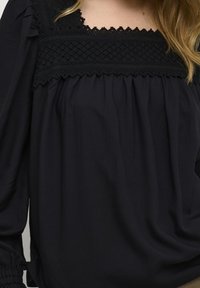 Blusa nera con yoke in pizzo texturizzato e scollatura arricciata. Maniche lunghe con polsini elastici, tessuto morbido con design drappeggiato.