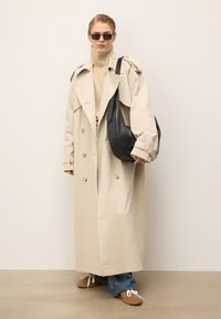 Osoba ubrana w beżowy trencz, brązowy golf, niebieskie dżinsy, brązowe buty, czarną torbę na ramię i ciemne okulary przeciwsłoneczne, stojąca przy jasnej ścianie.