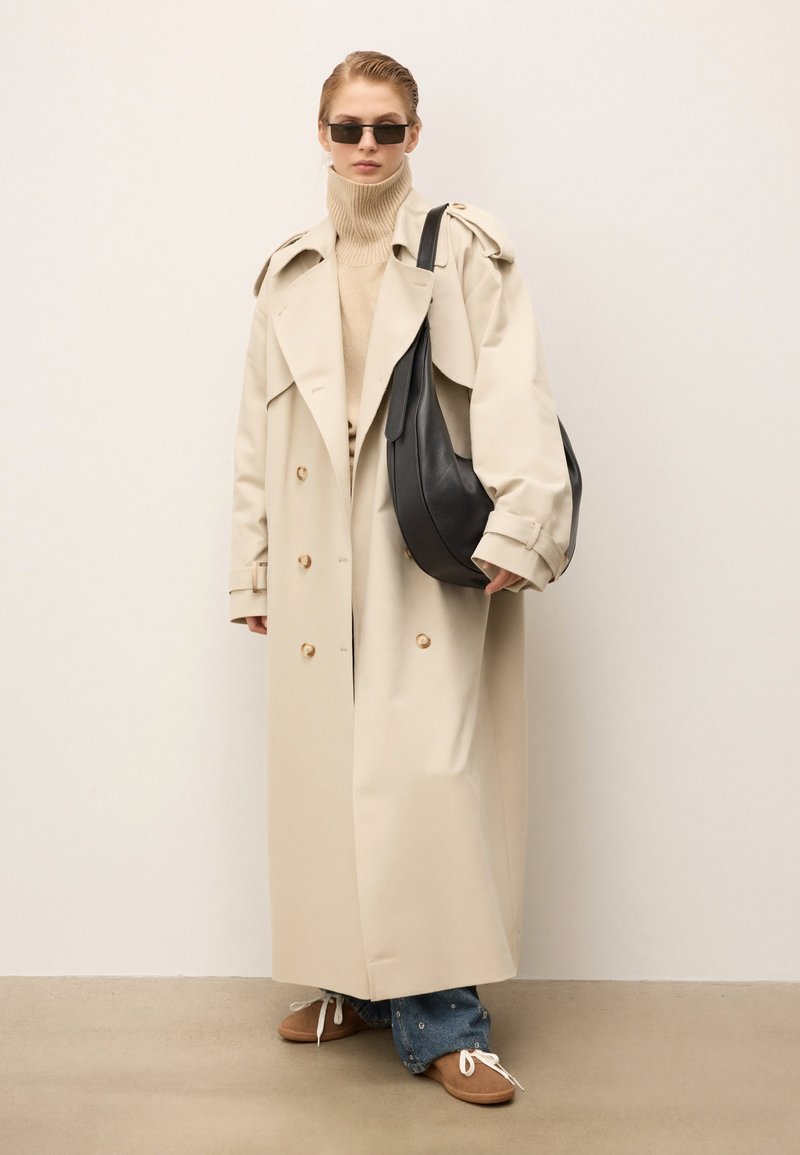 Osoba ubrana w beżowy trencz, brązowy golf, niebieskie dżinsy, brązowe buty, czarną torbę na ramię i ciemne okulary przeciwsłoneczne, stojąca przy jasnej ścianie.