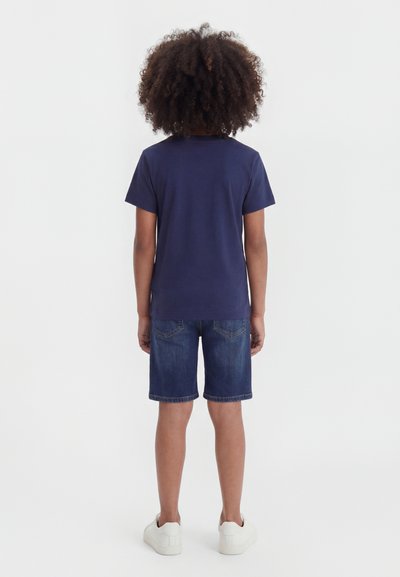 Enfant debout de dos, portant un t-shirt bleu marine, un short en jean et des baskets blanches sur un fond blanc uni.