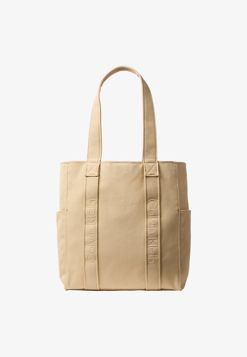 Sac cabas en toile beige Calvin Klein avec deux longues anses, broderie verticales du nom de la marque sur les bretelles avant, et poches latérales.