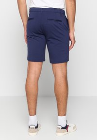 Man wearing navy blue tailored shorts, white crew socks, and white sneakers standing facing away against a plain background.