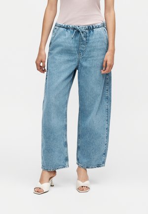 Person iført lyseblå denim-bukser med brede ben og snøreliv, hvide sandaler med hæl og en lyserød top, står mod en hvid baggrund.