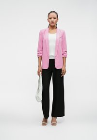 Ružový prispôsobený blejzer, biele tričko, čierne široké nohavice a béžové podpätky; drží bielu tašku s perforovanými detailmi. Jednoduché pozadie.